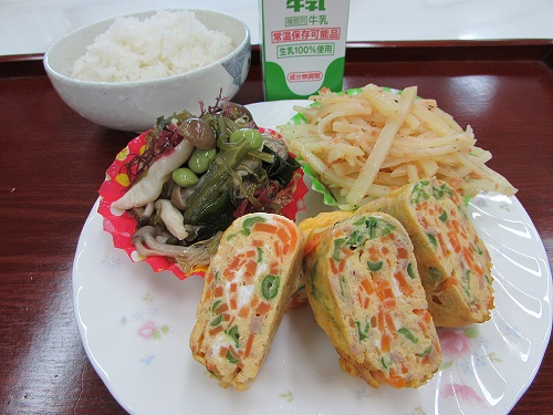 お皿の上にニンジンなどが入った卵焼き、しめじの海藻和え、じゃがいもと鮭フレーク炒めが載っており、その奥にはご飯と牛乳が置かれてある写真