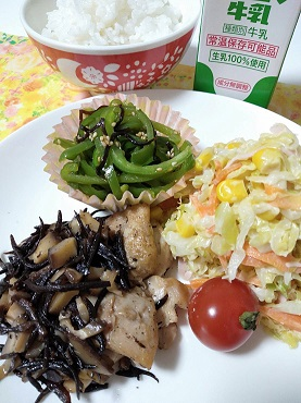 ひじきと干しシイタケの照り焼きチキンとコールスローサラダとおかずカップに乗ったピーマンの塩昆布和えが乗っている皿と、その奥に牛乳とご飯がある写真