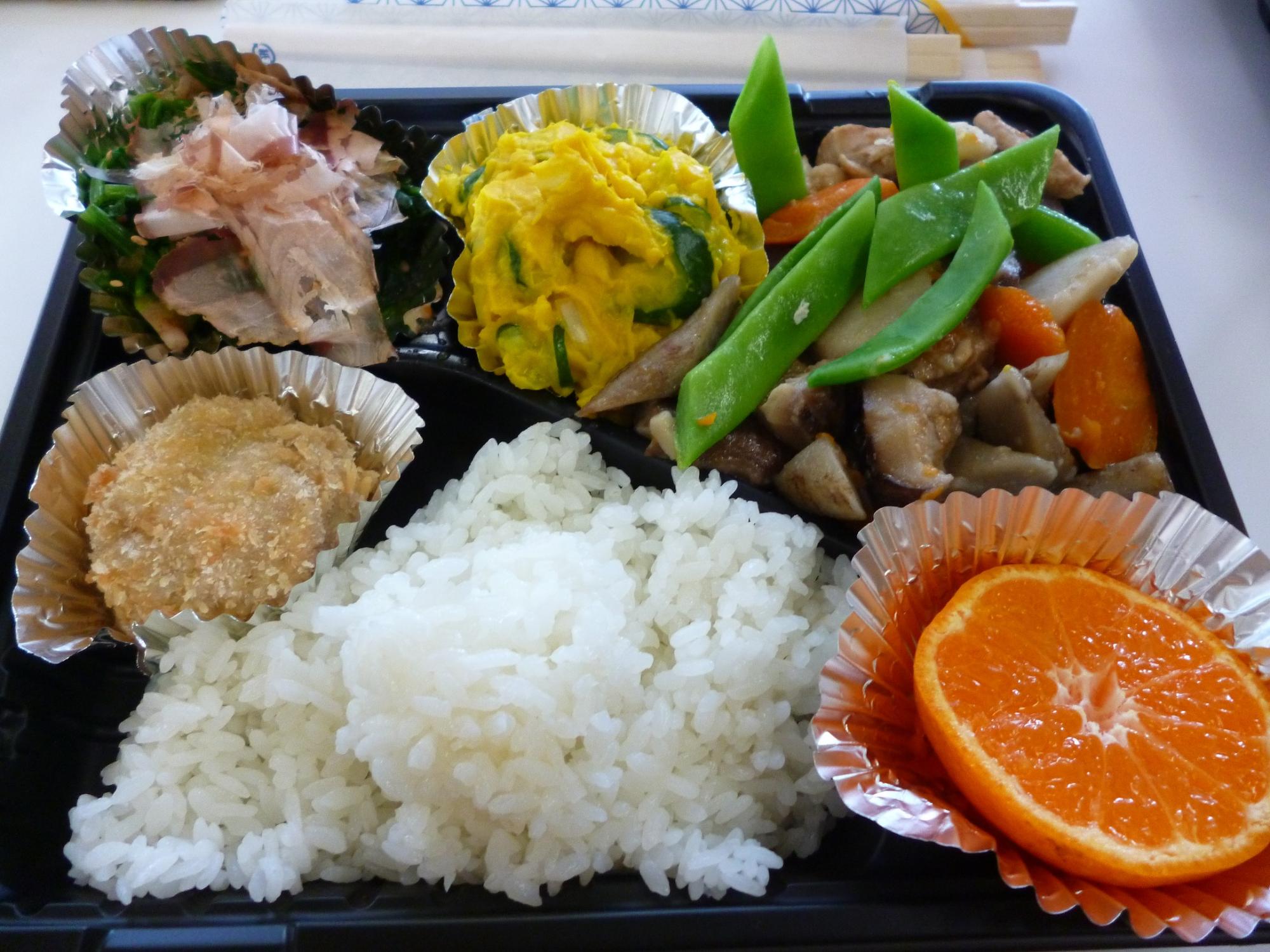 弁当容器に筑前煮、すり身揚げ、かぼちゃのサラダ、ほうれん草のゴマ味噌酢和え、ご飯、オレンジが盛り付けられた写真