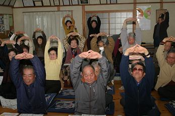 一室に集まった団体の方々が両手を組んで上に挙げている介護予防教室の写真