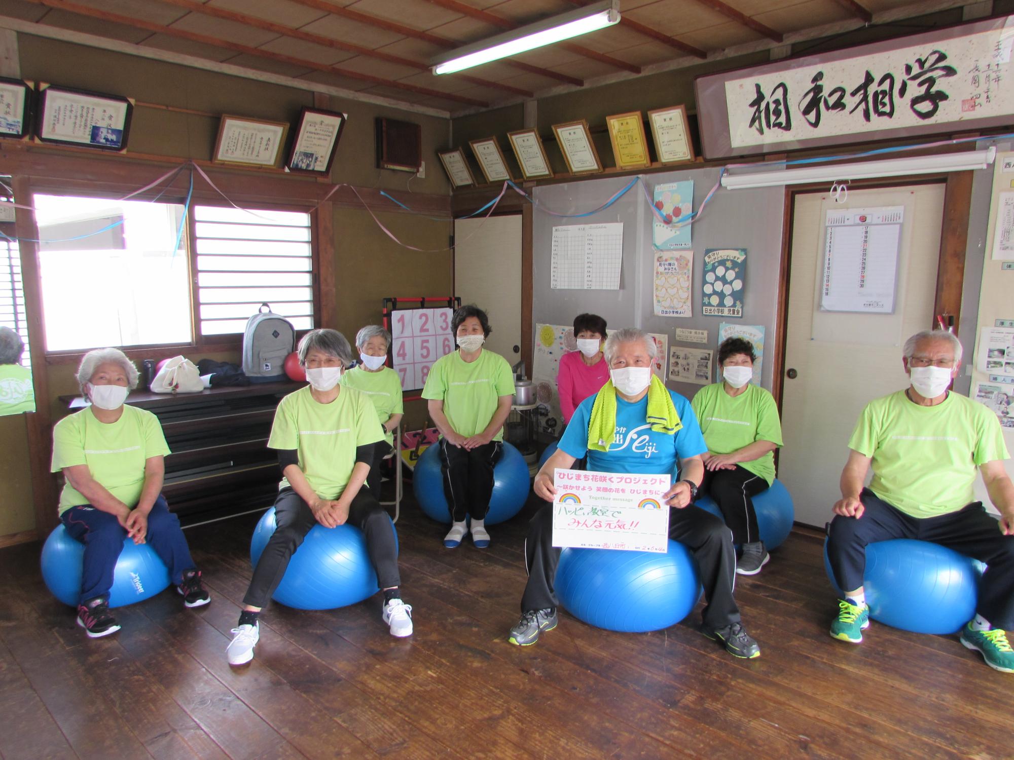 室内で2名が椅子に、6名が水色のバランスボールに座り、前右から2番目の男性が両手で応援メッセージの用紙を持っている集合写真