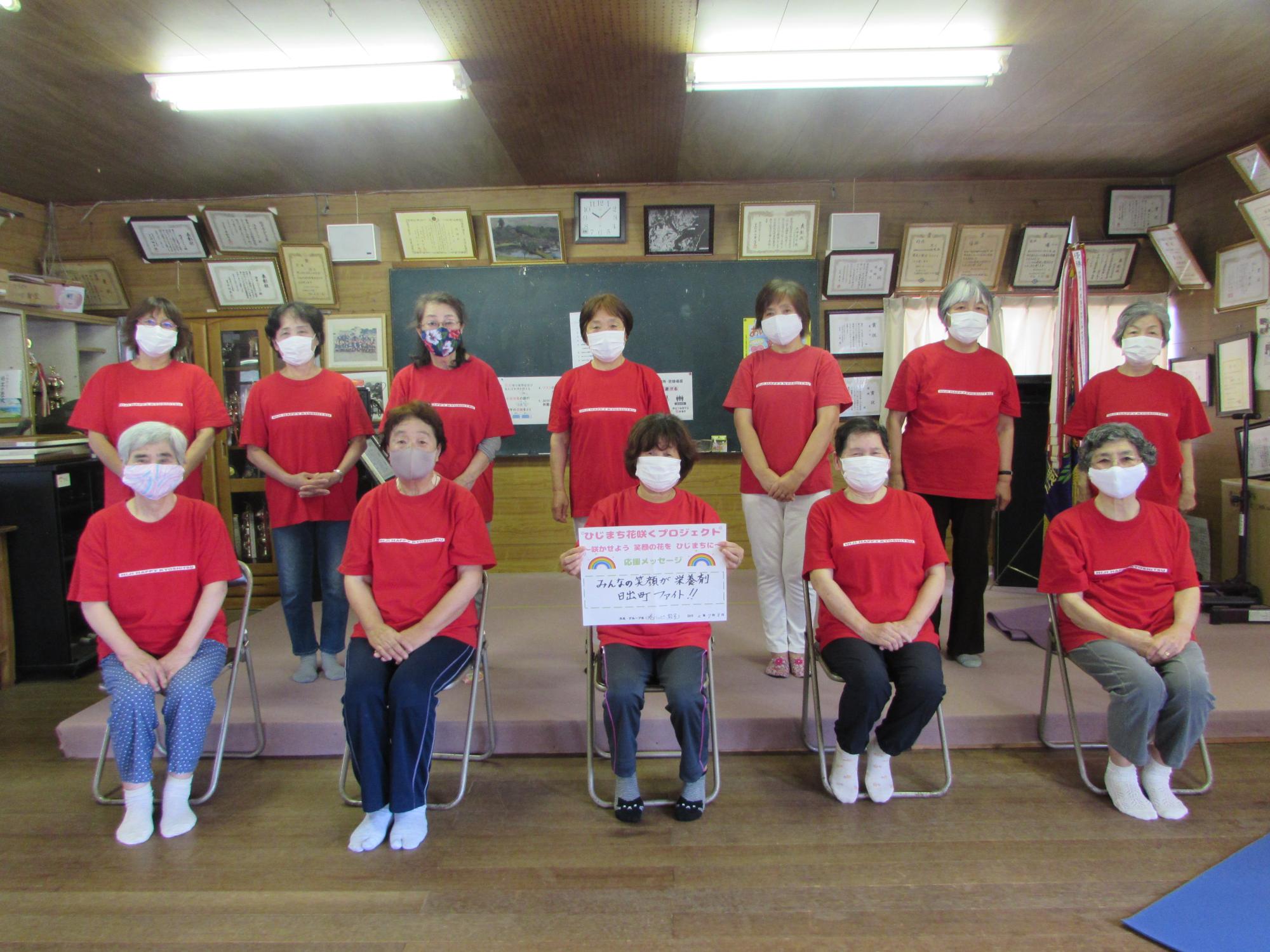 お揃いの赤いシャツを着た12名の女性たちが集まり、前中央のパイプ椅子に座っている女性が両手で応援メッセージを持っている集合写真