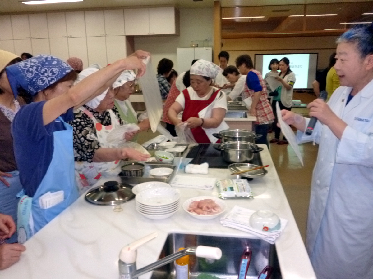 青い三角巾にエプロン姿の女性が調理台の反対側に立つ白衣の女性にポリ袋を持ち上げ見せ、周りでは沢山の参加者の人達が調理の準備をしている写真