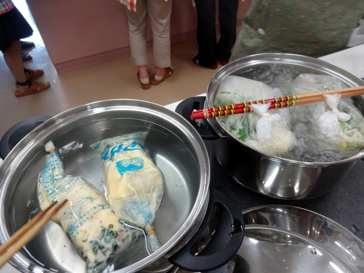 右の鍋の中に材料を入れた2つのポリ袋を温め、左の鍋ではポリ袋に入った材料を菜箸で押さえている写真