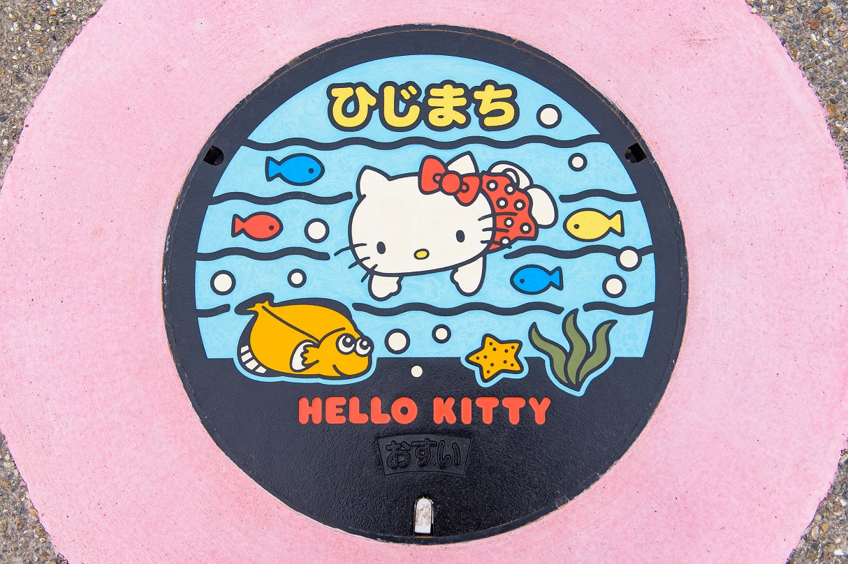 ハローキティがカラフルな魚たちと水中で泳ぐ様子が描かれ、ひじまち HELLO KITTYと印字されたマンホール