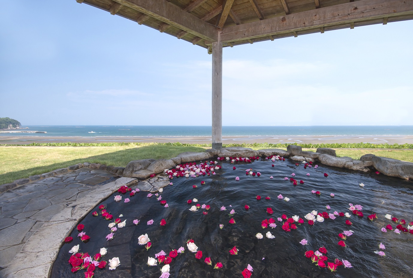 赤・白・ピンク色の花が浮かべられた円形の石造りの温泉と青い別府湾を間近に望む露天風呂の様子