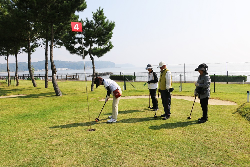 男女4人がゴルフクラブを手に立っており、その内1人がパターを決めようとしている写真