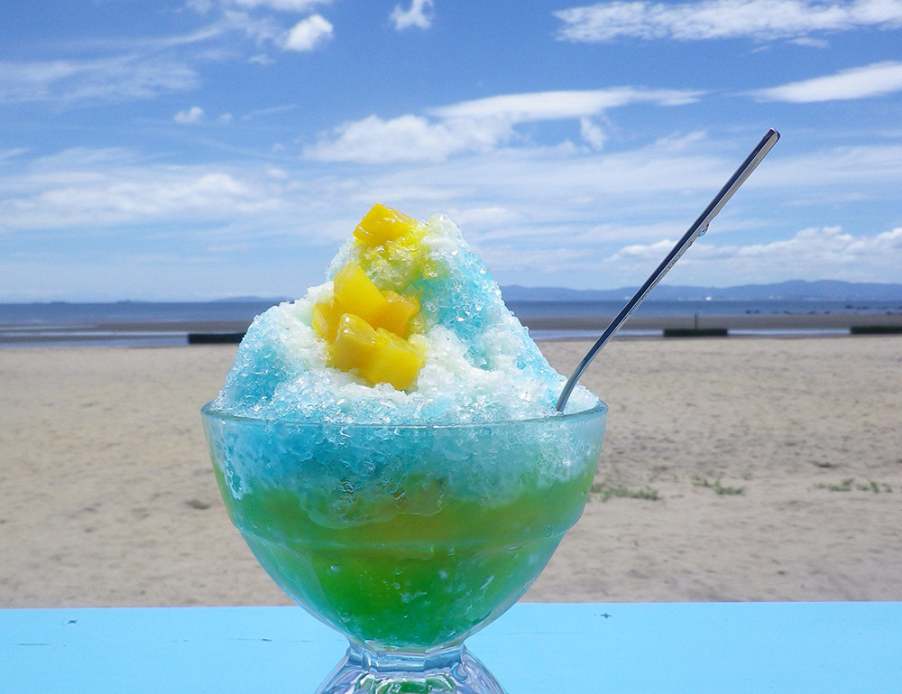 奥に海と空が広がる砂浜で手前の机に青いソースと黄色いフルーツのかかったかき氷の写っている写真