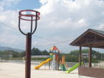 奥に滑り台と木造の休憩所、手前にバスケットボールのゴールのような鉄製のボール入れが写っている写真