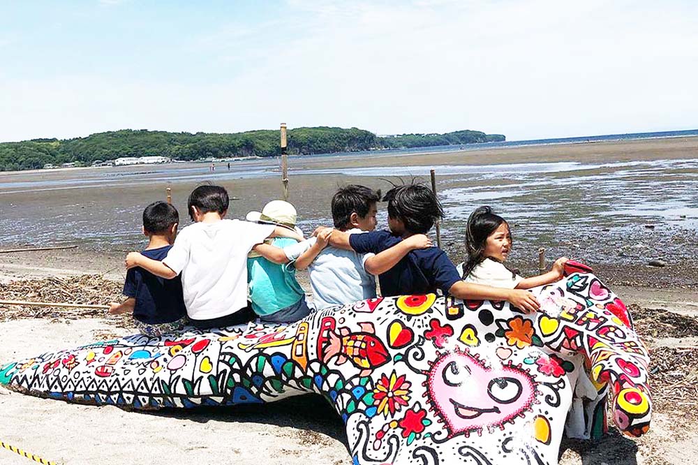 海岸のそばに置かれた糸ヶ浜に流れ着いた流木に、ハートや太陽、花など色とりどりなアーティスティックなペイントを施したベンチに数人の子供たちが背中を向けて肩を組みながら座っている写真