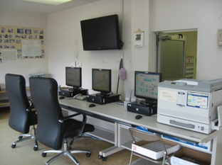 手前に背もたれのある椅子が2つあり、その前の机に左からパソコンとモニターが3台あり、その右にプリンターが置いてある写真