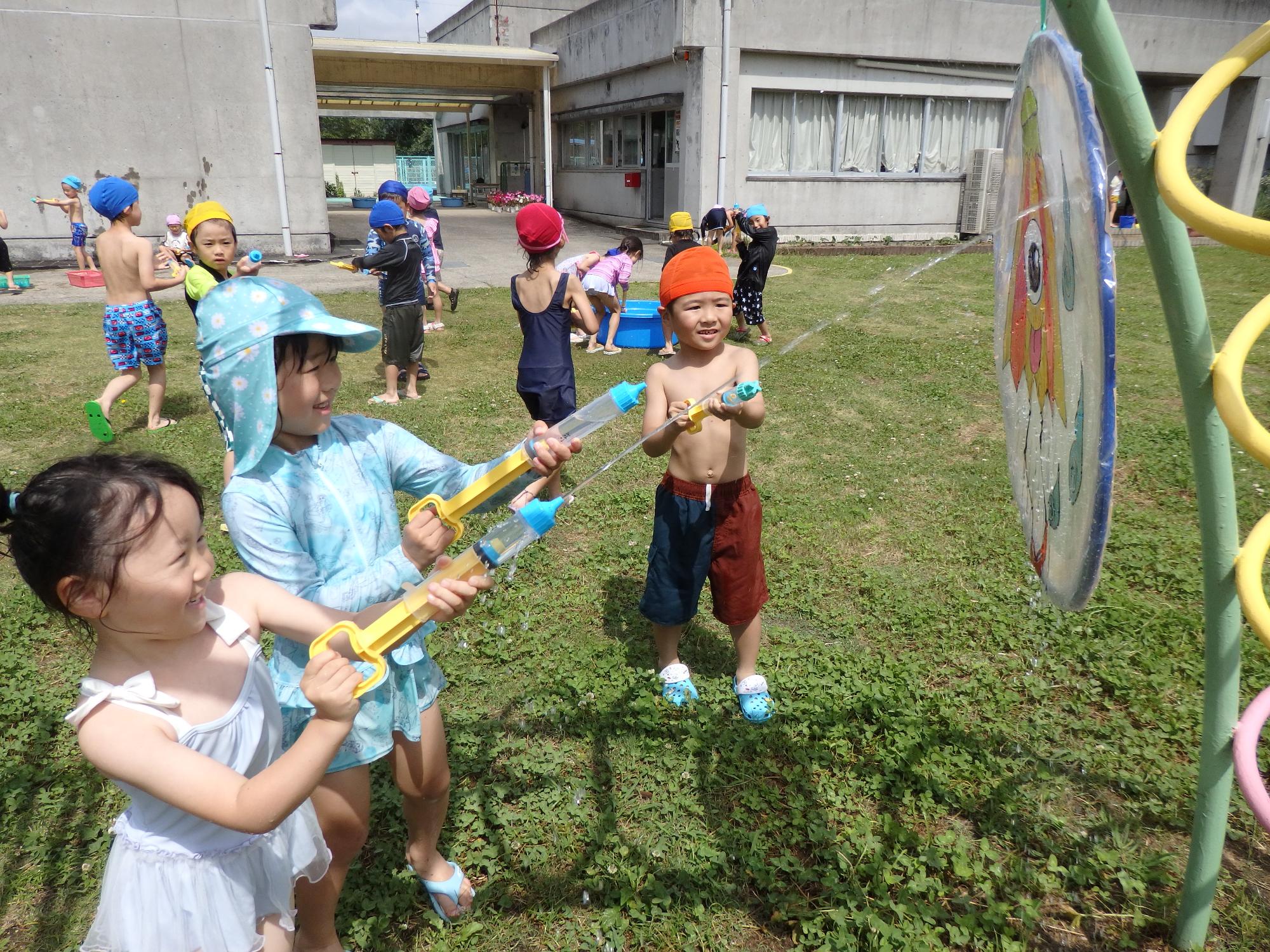 園庭で水着の園児達が水鉄砲を持って、的に水を当てる様子の写真