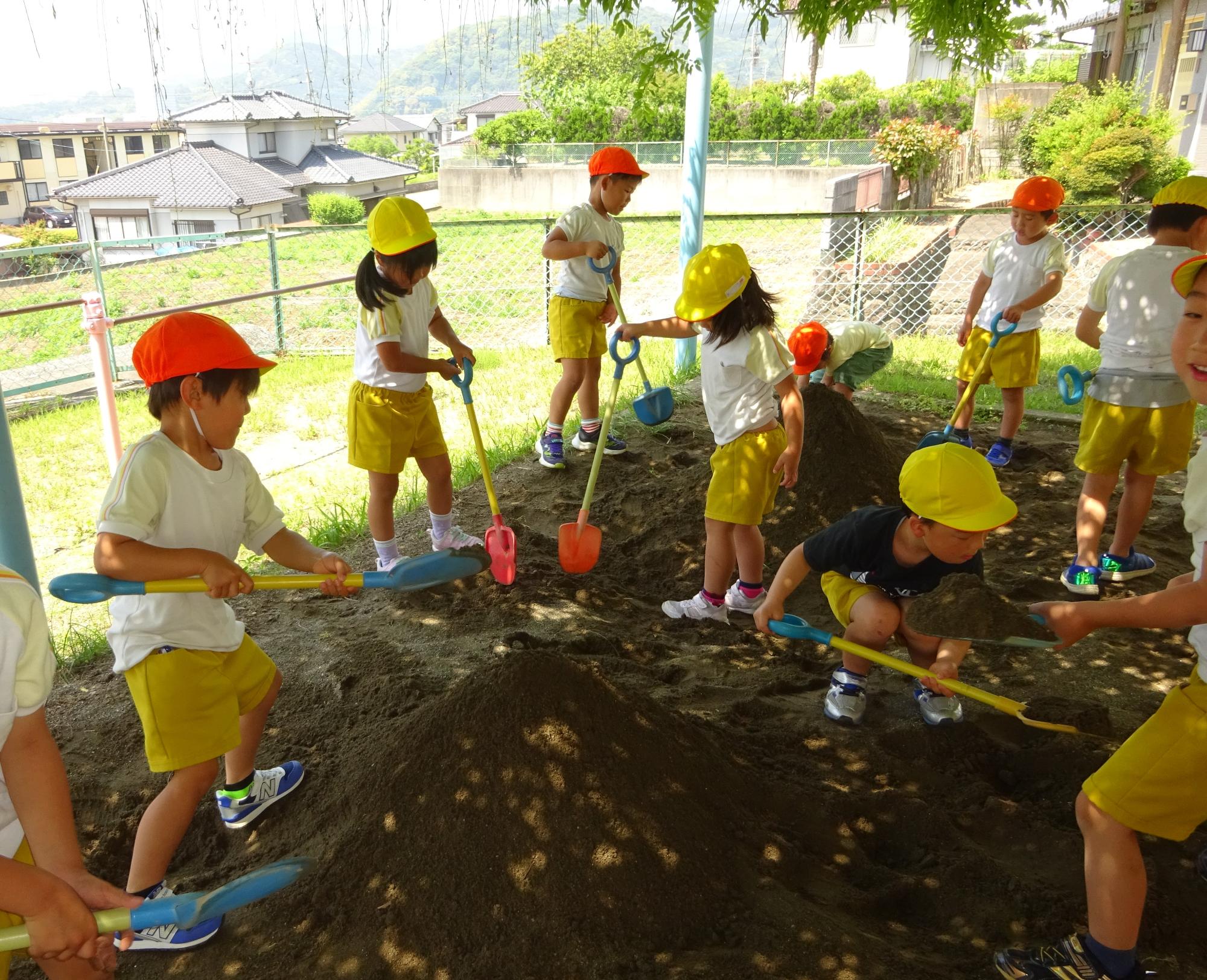オレンジと黄色の帽子をかぶった園児9人が砂場であそんでいる