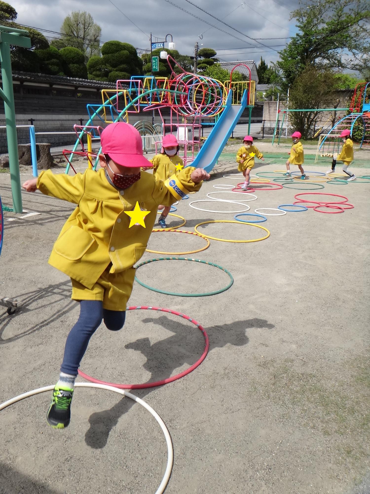 園庭で赤い帽子を被った園児たちが、カラフルなフープを次々に跳んでいる様子の写真