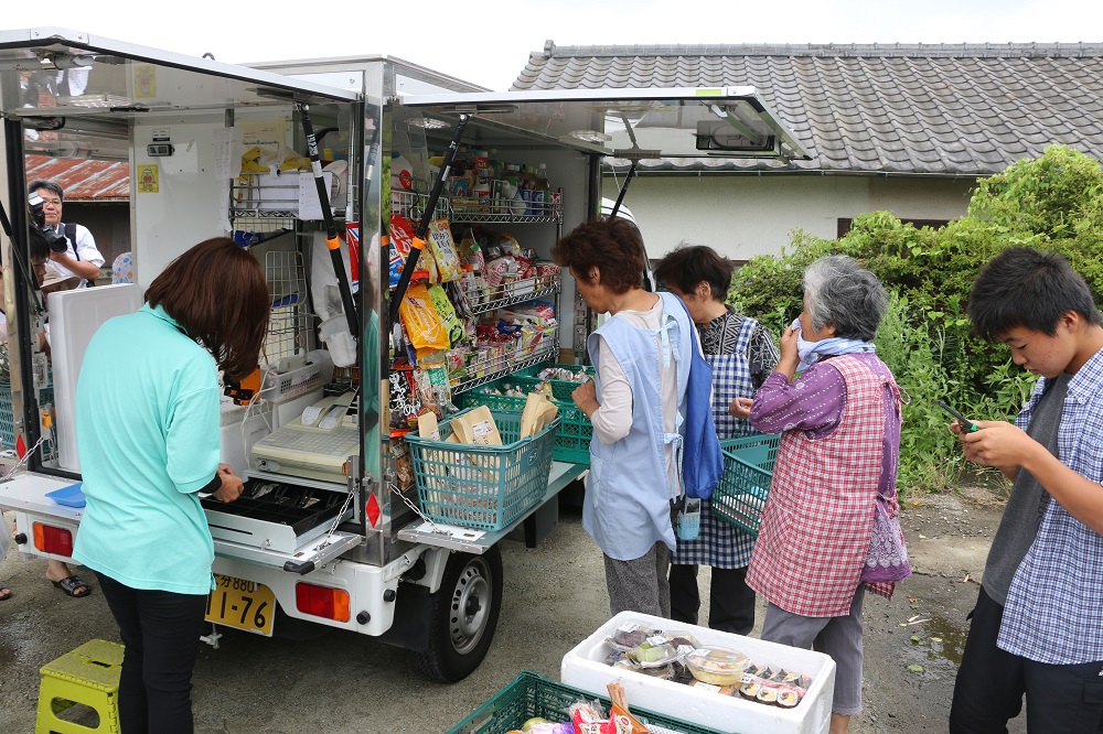 移動販売車の荷台が広げられ、そこに陳列された食料品などの商品を見ている3人の女性客とレジの前に立つ販売員の後姿を撮った写真